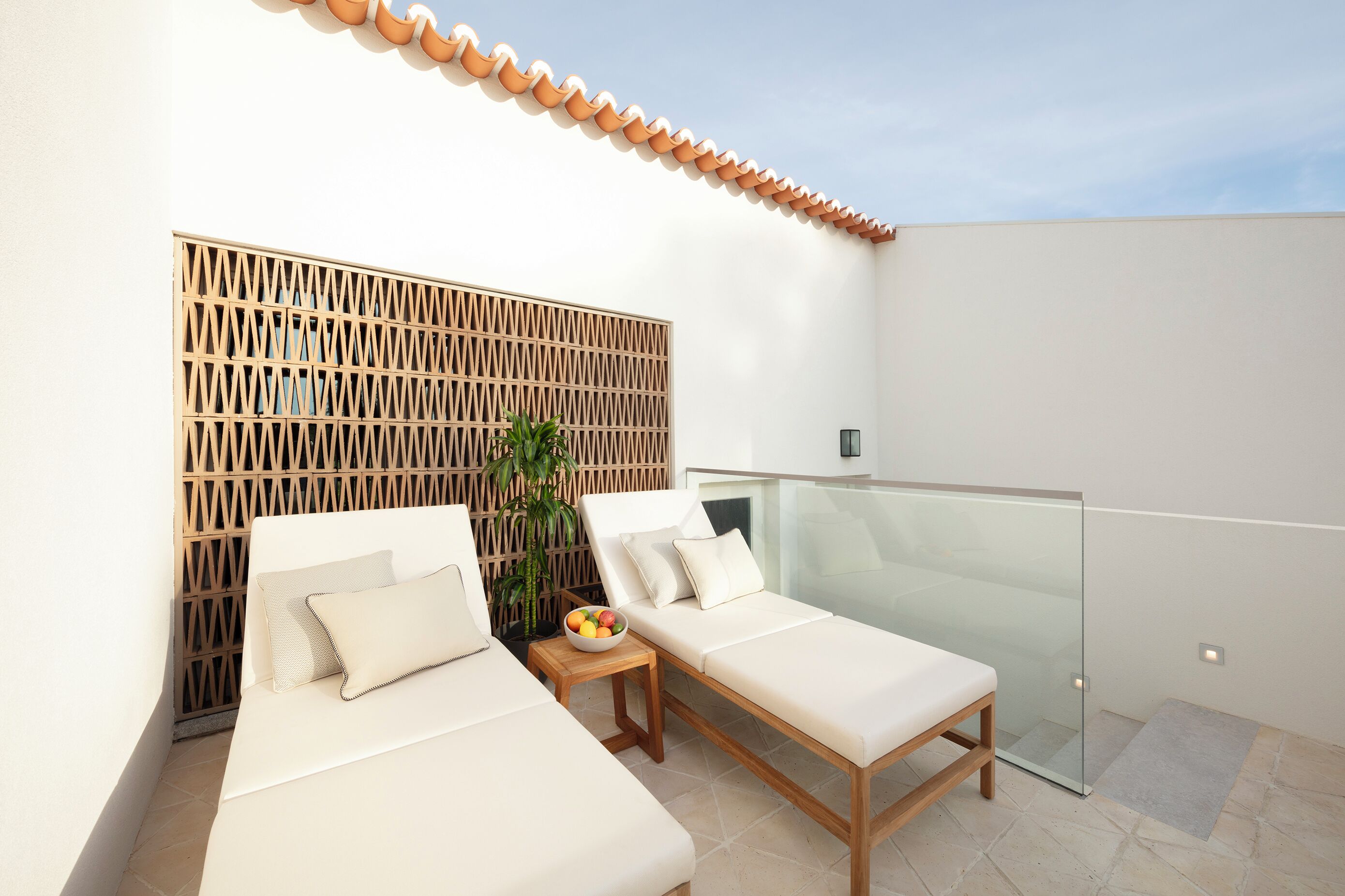 A balcony with two white lounge chairs, a wooden table with a bowl of fruit, and a potted plant.