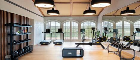 A spacious gym with treadmills, elliptical machines, and weight equipment. Large windows provide natural light and views of the outdoors.
