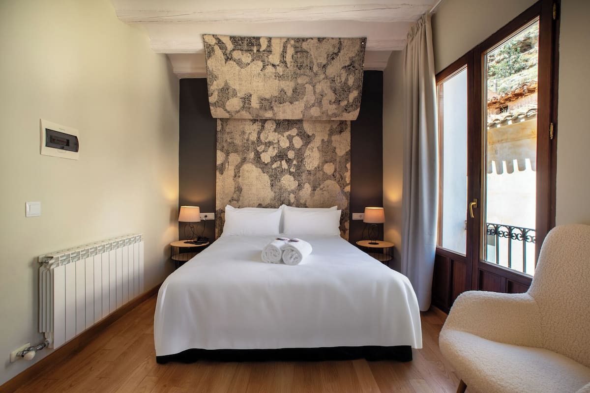 Habitación de hotel con una cama grande, dos mesitas de noche con lámparas, un radiador y vistas a un edificio a través de una ventana.