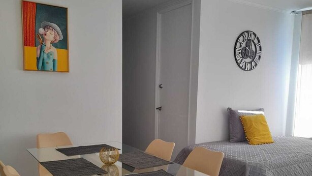 Moderni huone, jossa on lasinen ruokapöytä, beigenväriset tuolit, harmaan- ja keltaisen värinen päiväpeite, seinäkello ja taulu.