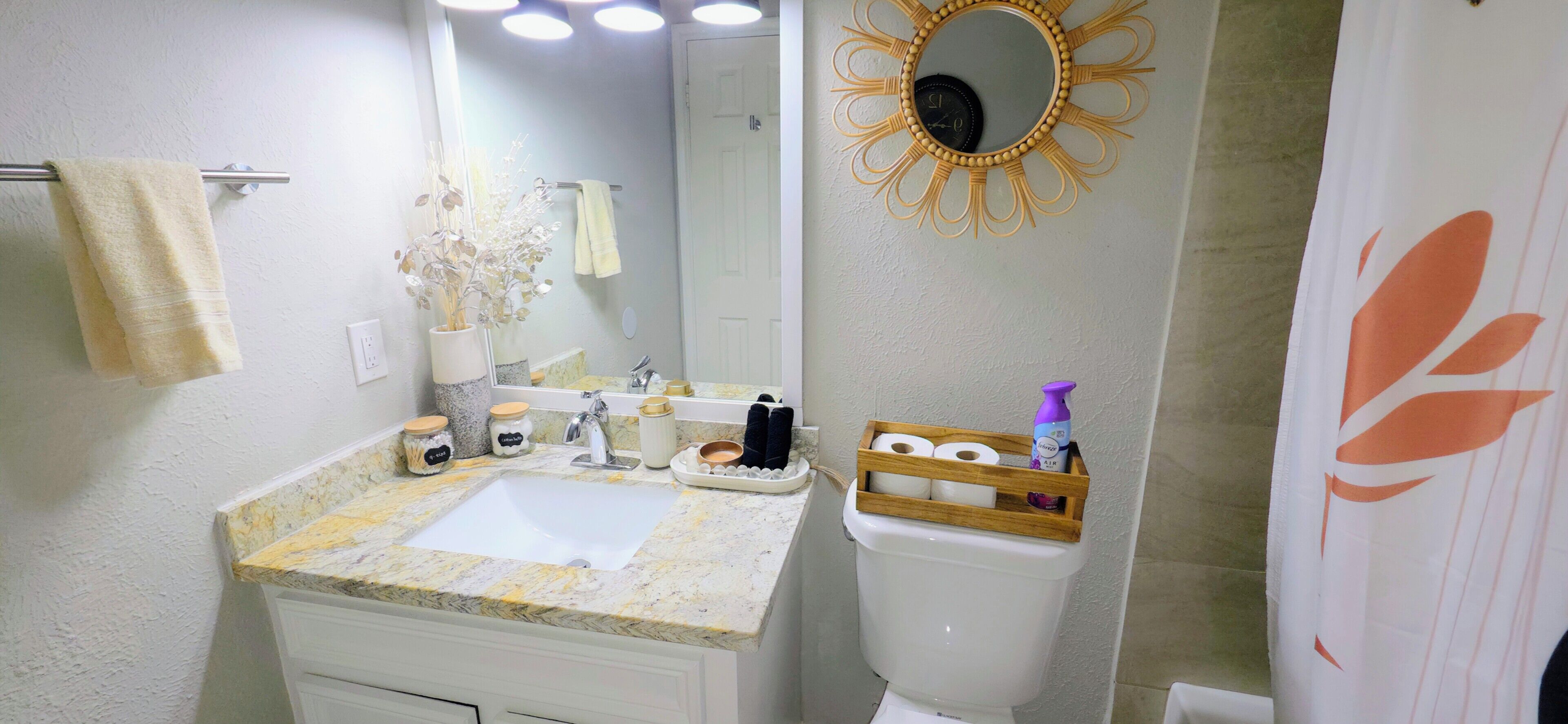 A bathroom with a sink, mirror, and toilet. There are towels hanging on the wall, a decorative mirror frame, and a shower curtain with a floral design.