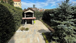 A paved pathway leading to a building with a red roof and a chimney, surrounded by dense greenery and a clear blue sky.