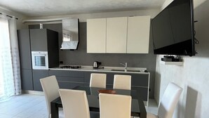 A modern kitchen with a dining area, featuring a central island, white chairs, and a flat-screen TV mounted on the wall.