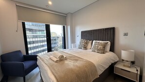 Et moderne hotelværelse med en stor seng, en stol, et natbord med lampe og udsigt over byen gennem et stort vindue.