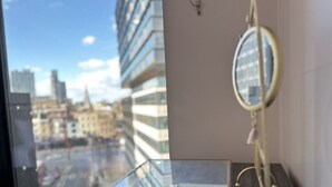Et værelse med udsigt til bybilledet gennem et vindue, et natbord med et spejl, og et lille natbord med dekorative genstande.