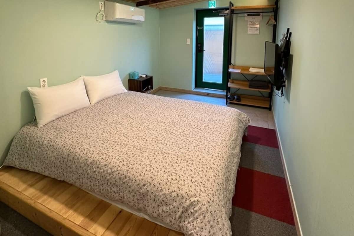 A bedroom with a wooden bed, a small bedside table, a nightstand, a window with a view, and a door leading to another room.