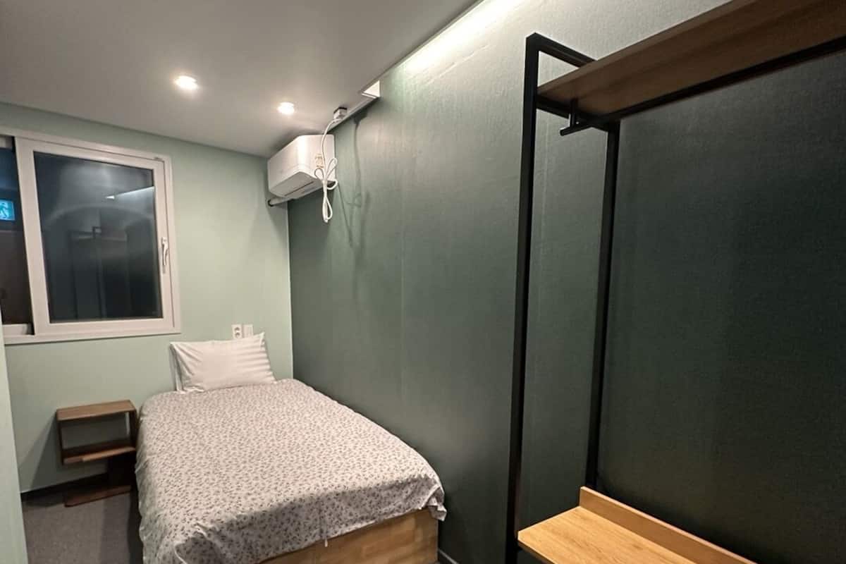 A bedroom with a bed, a window, a bedside table, and a wall-mounted shelf.