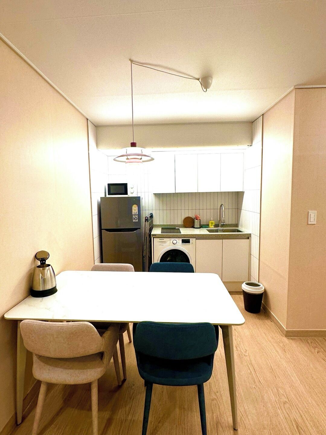A compact kitchen with a refrigerator, microwave, washing machine, and a small countertop. There is a dining table with two chairs and a hanging light fixture above it.
