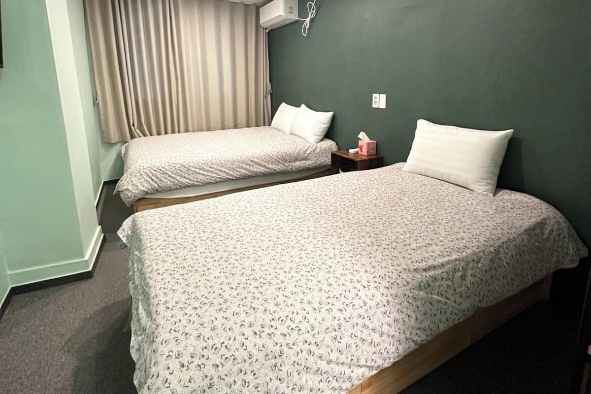 A room with two single beds, a small bedside table with a lamp, a wall-mounted air conditioner, and a window with curtains.