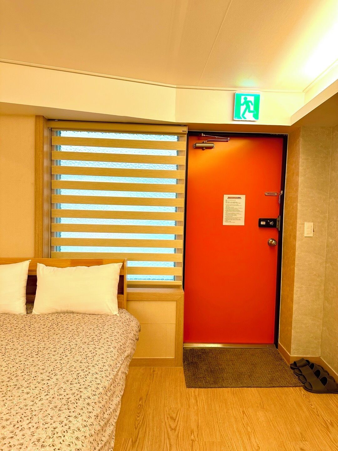 Una habitación de hotel con una cama, una ventana con persianas, una puerta naranja y un felpudo pequeño en la entrada.