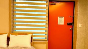 Una habitación de hotel con una cama, una ventana con persianas, una puerta naranja y un felpudo pequeño en la entrada.