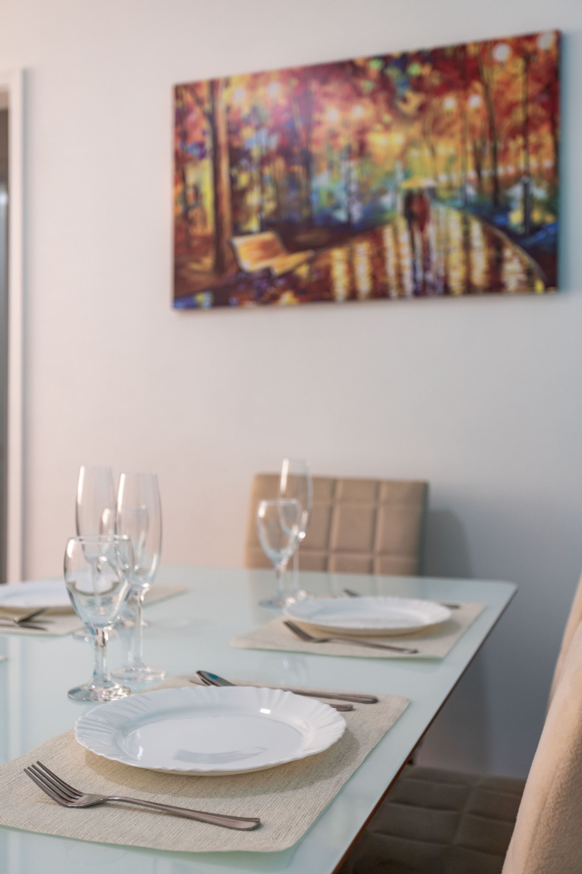 A dining table set with plates, cutlery, and wine glasses, with a colorful painting on the wall behind.