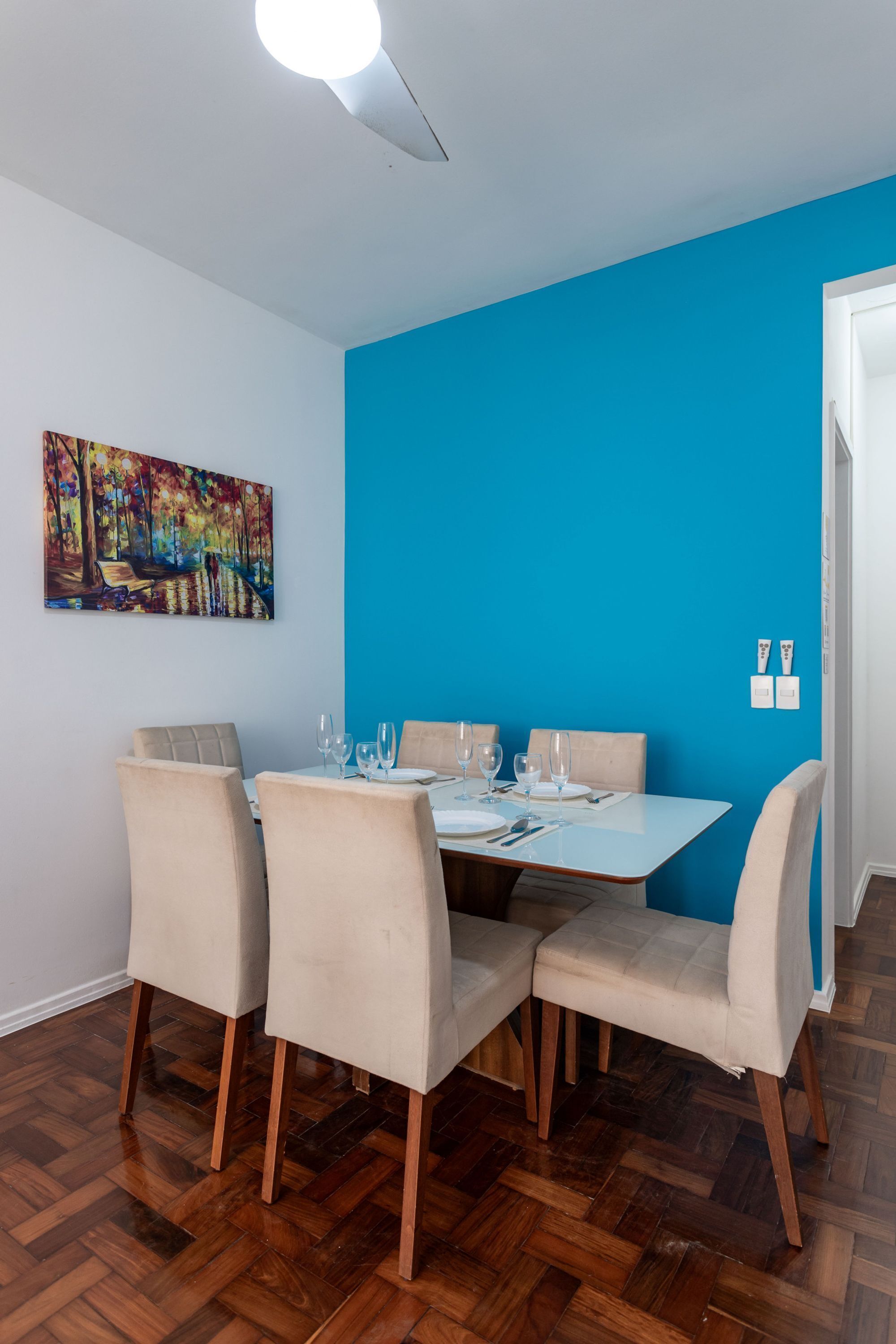 Comedor con mesa blanca para cuatro, sillas color beige, pared de acento azul, piso de madera y una lámpara en el techo.