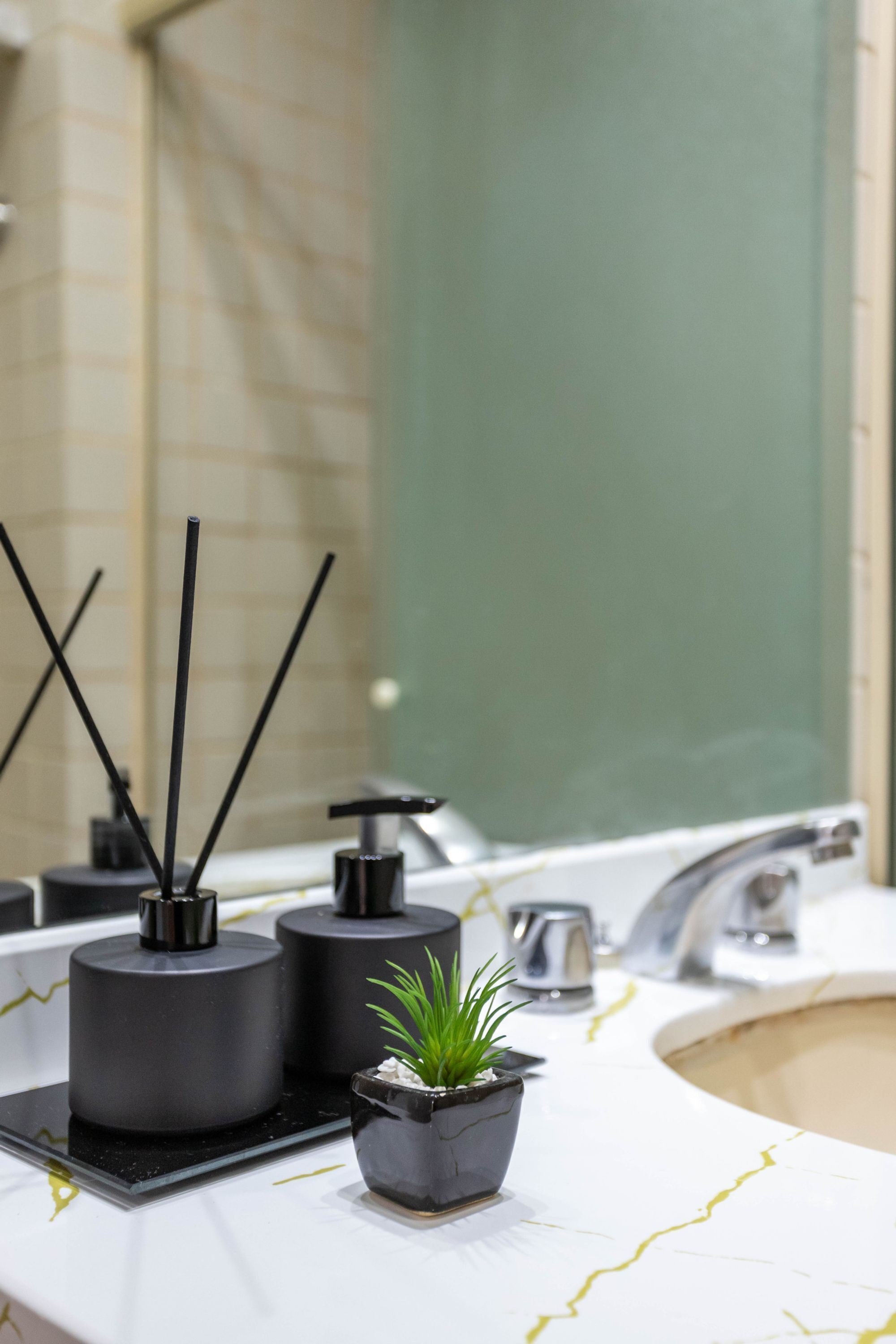 Un baño con lavamanos de mármol blanco, dispensadores de jabón negros, una pequeña planta en maceta y un espejo.