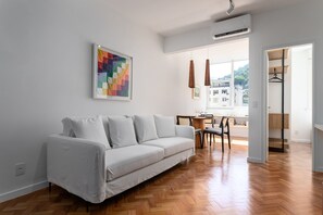 A modern living room with a white sofa, wooden herringbone flooring, a dining area with a table and chairs, and a view through a large window. - 5 Minutes From the Beach and a View of Christ Br904 (Rio de Janeiro)