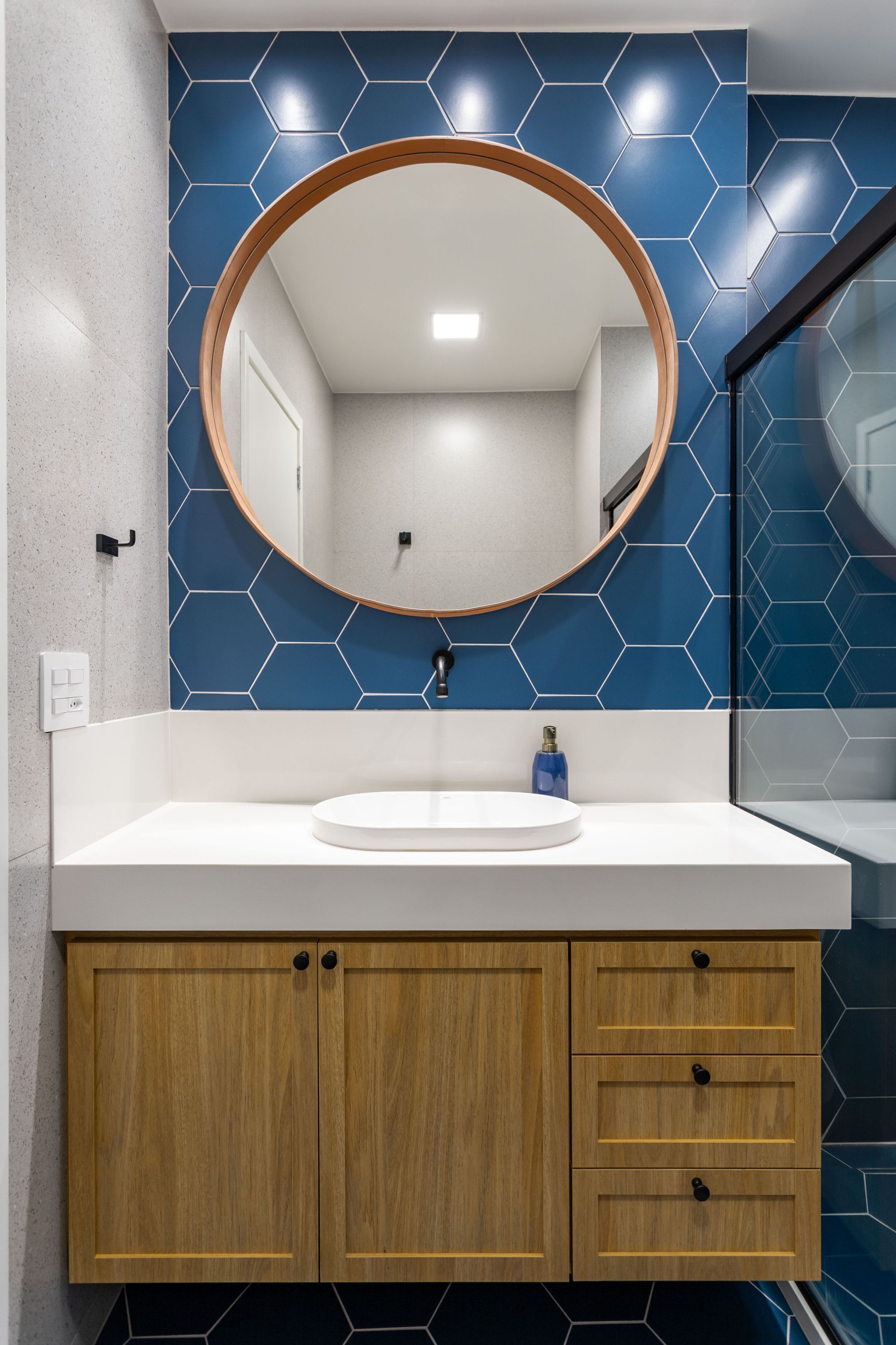 A bathroom with a round mirror, a white sink, wooden cabinets, and blue hexagonal tiles on the wall.