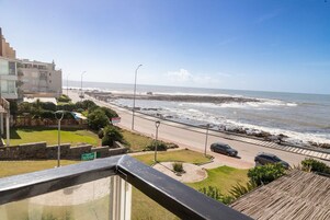 Vista costera desde un balcón con vista a una playa, con una carretera, coches y edificios al fondo.