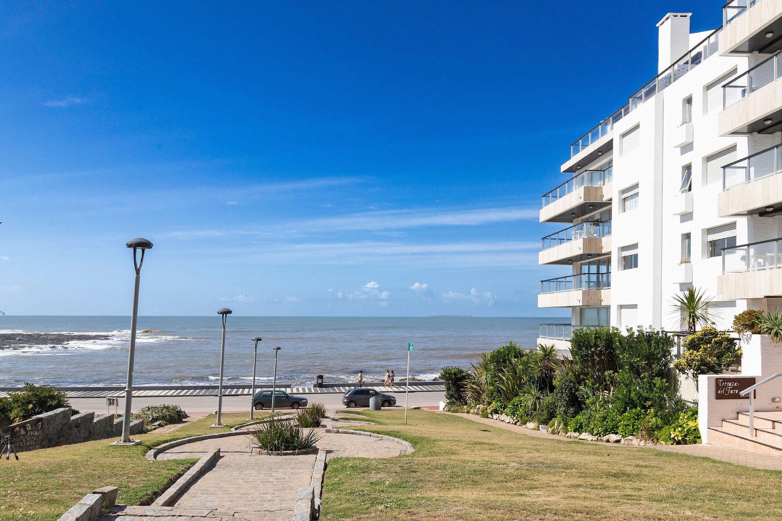 Un bâtiment situé sur la côte, doté de balcons donnant sur la mer, d’une promenade pavée et d’une zone herbeuse.