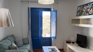 Una sala de estar con una puerta azul, muebles blancos, un televisor y un cuadro en la pared.
