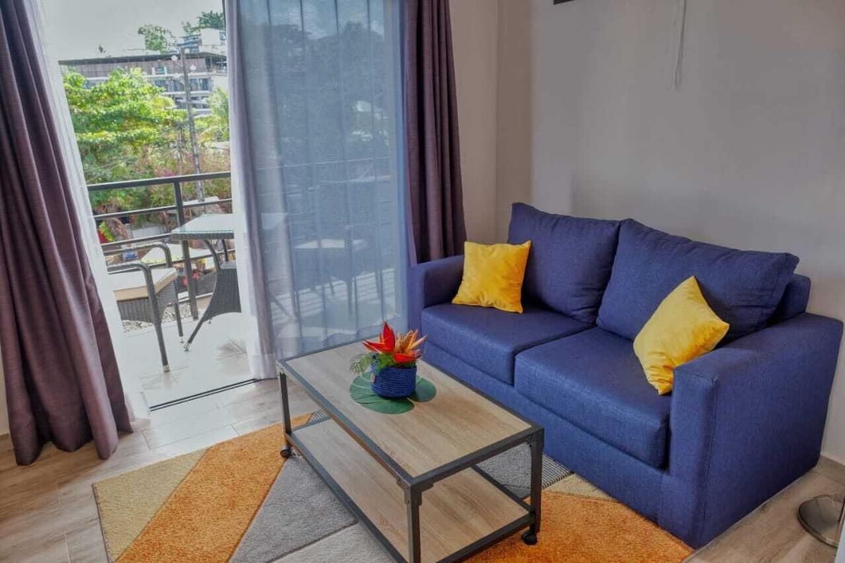 A modern living room with a blue sofa, yellow cushions, a glass coffee table, and a view of a balcony with outdoor furniture.