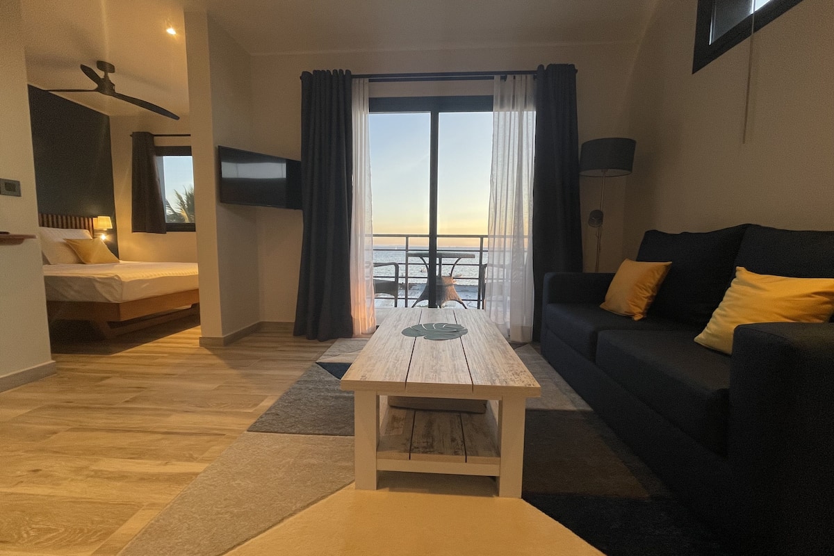 A modern living room with a sofa, a coffee table, a bed, and a balcony with a view of the sea.