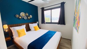 A hotel room with a bed, a nightstand, a wall clock, a window with curtains, and a view of the outdoors. - BLUE VELVET (Nosy Be)