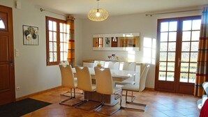 A dining area with a white table and chairs, a hanging light fixture, and large windows with curtains. - Le Gîte Isabel en Dordogne, Près de Sarlat (Saint-Crépin-et-Carlucet)