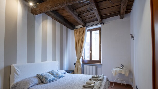 A bedroom with a bed, a window with curtains, a small table with a lamp, and a radiator.