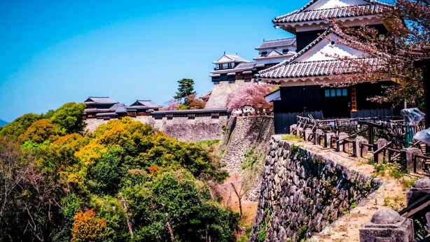 A traditional Japanese castle with multiple buildings, a stone wall, and a forested hillside. - Himerakuen媛楽園　松山市中心部にある家族・グループ旅行の多人数が泊まれる最適な宿 (松山市)