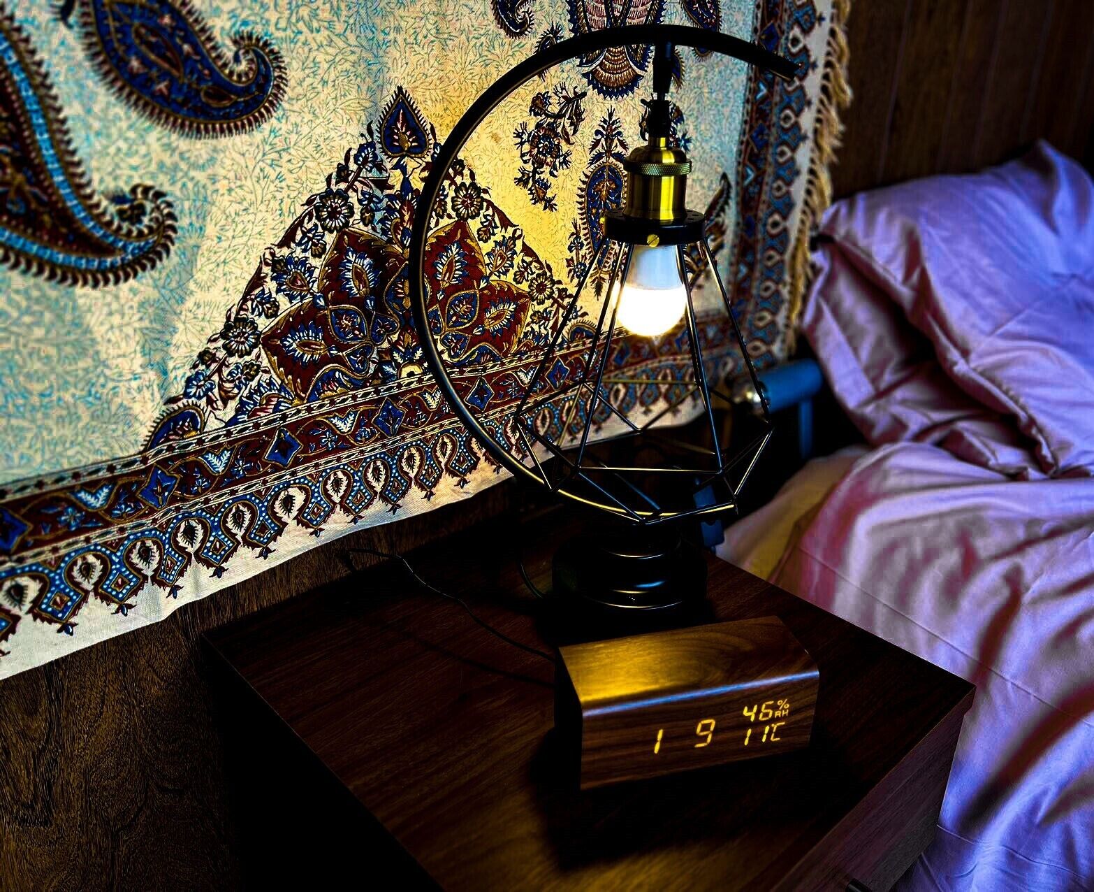 A bed with a wooden bedside table, a lamp, and a digital clock displaying 19:11.