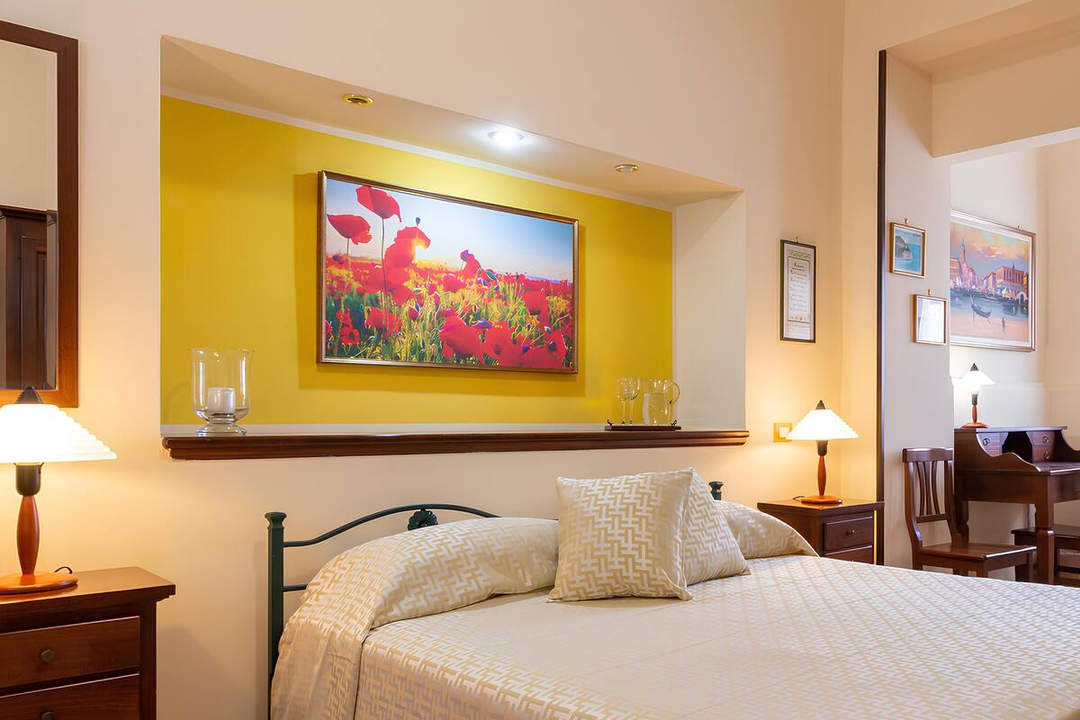 Habitación de hotel con cama, dos mesitas de noche, un escritorio y una silla. Hay un cuadro en la pared sobre la cama y una fotografía enmarcada a la derecha.