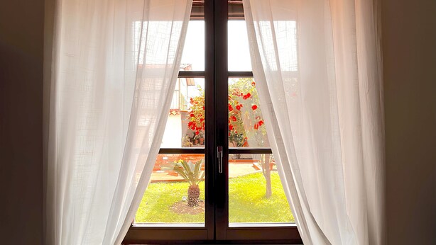 A window with white curtains, a view of a garden with red flowers, and a wooden floor. - B&B Hibiscus (Agropoli)
