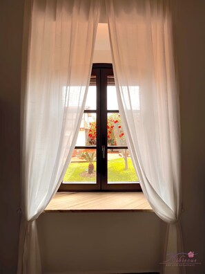 A window with white curtains, a view of a garden with red flowers, and a wooden floor. - B&B Hibiscus (Agropoli)