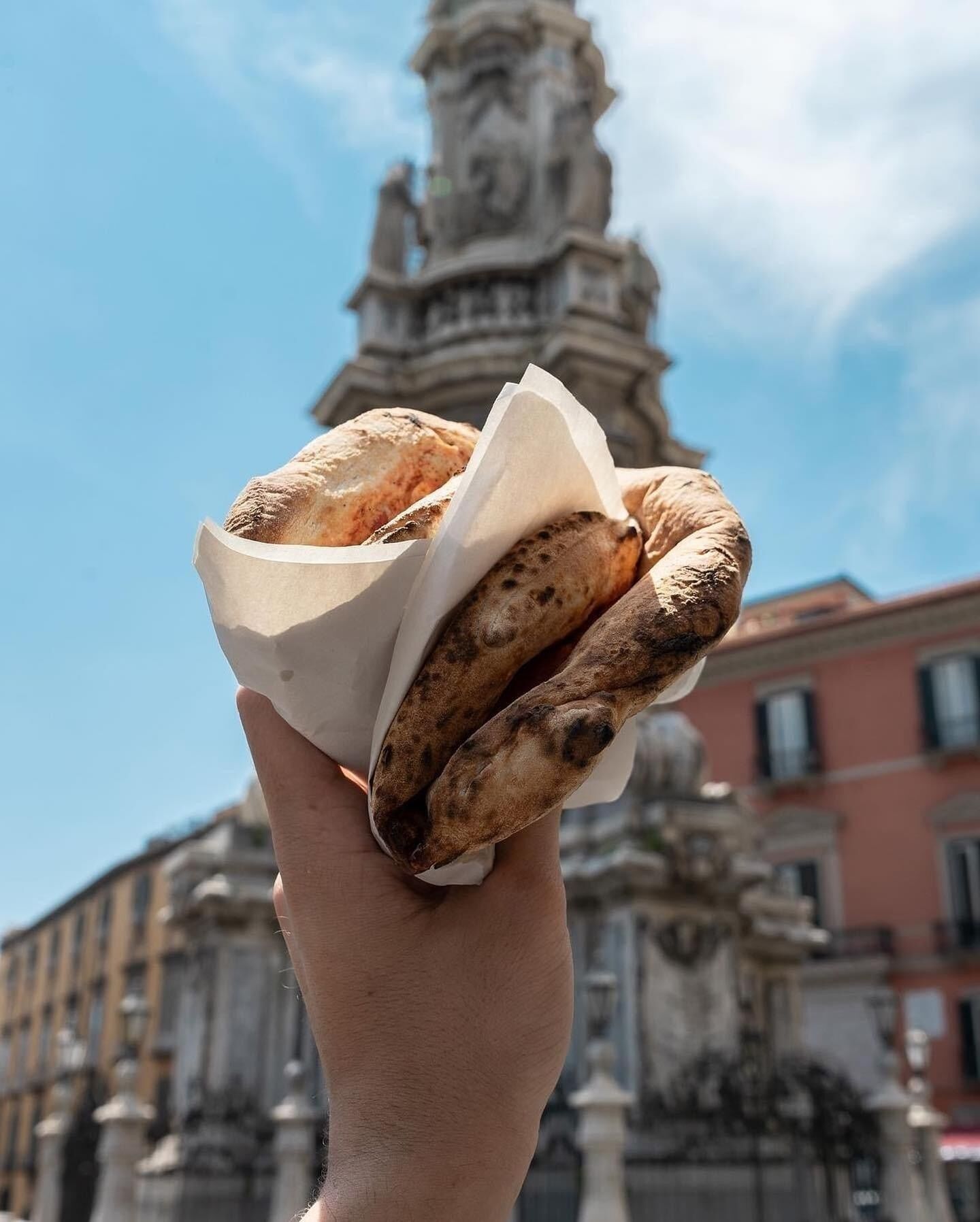 A hand holding a piece of flatbread with a paper wrapper, with a historic building and a statue in the background.