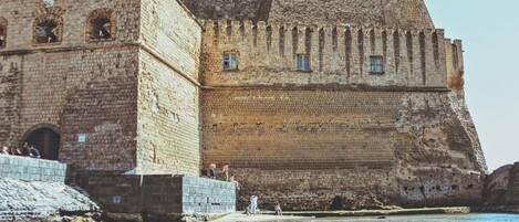 Una fortaleza histórica de piedra con una gran torre central y un muro fortificado, ubicada junto a una masa de agua.