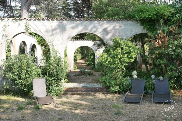 Un jardín con un arco de piedra, dos sillas de descanso y un camino que lo atraviesa.