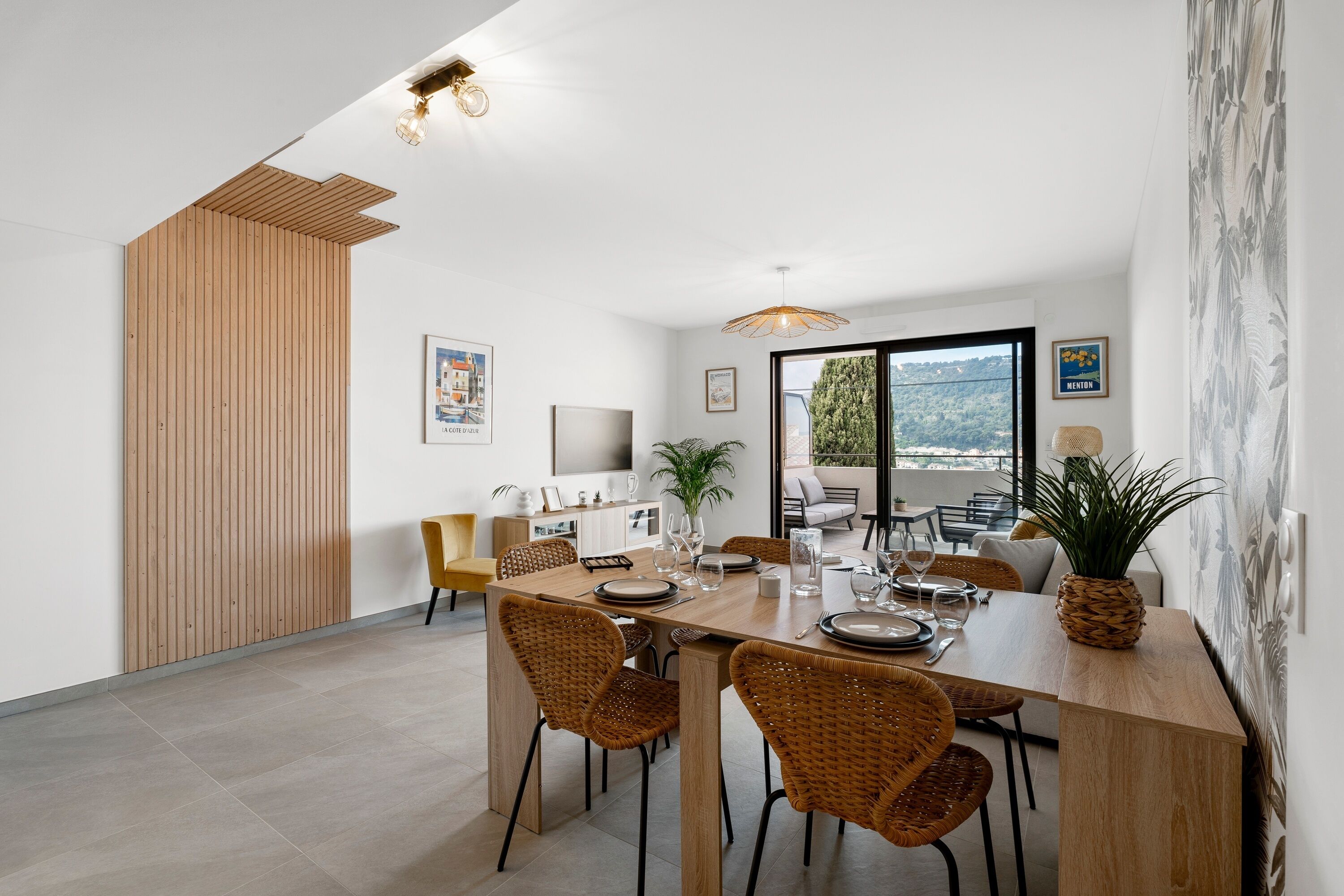 A modern dining area with a wooden table set for four, surrounded by wicker chairs. A large window offers a view of greenery outside.
