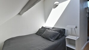 Un dormitorio moderno con techo inclinado, cama con ropa de cama gris, mesita de noche blanca y una lámpara fijada a la pared.