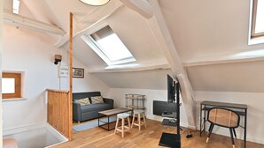 Un apartamento estilo loft con techo inclinado, tragaluz, sofá gris, mesa de centro de madera, dos taburetes, una silla negra y un mueble de TV negro.