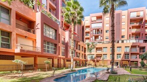 Un hotel de varios pisos con una piscina central, rodeado de palmeras y un césped bien cuidado.