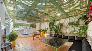 Un patio iluminado por el sol con una pérgola verde, una mesa de metal negro, un sofá negro y diversas plantas en macetas.