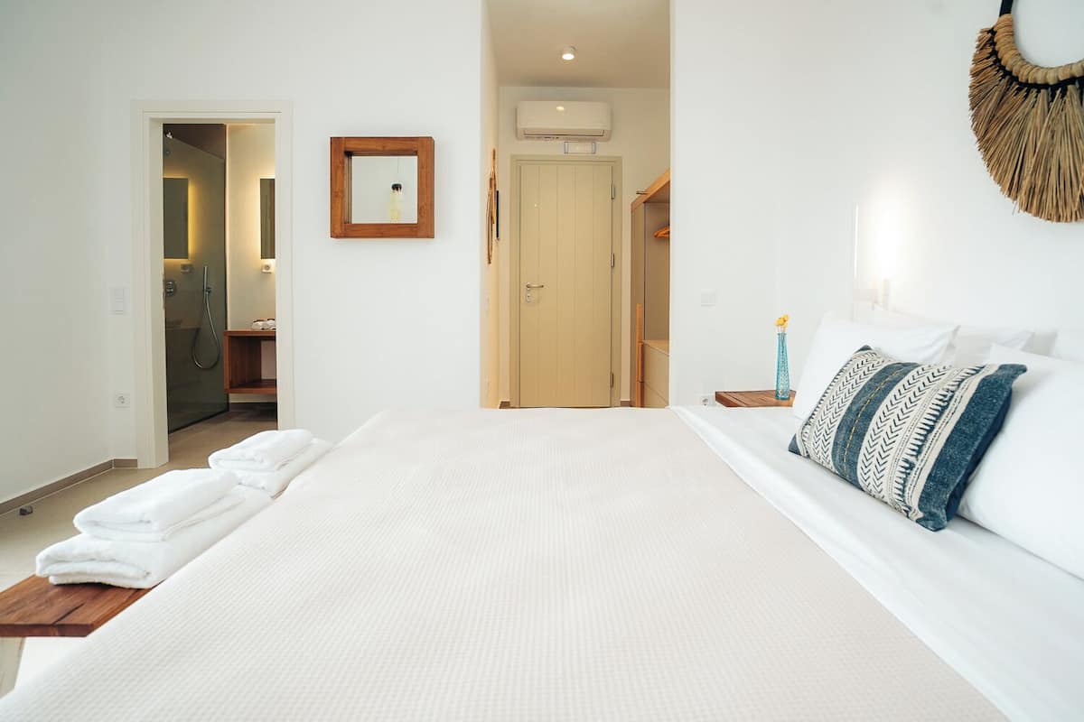 Quarto de hotel com uma cama grande, uma mesinha de cabeceira de madeira, um espelho e uma porta que dá para o banheiro.