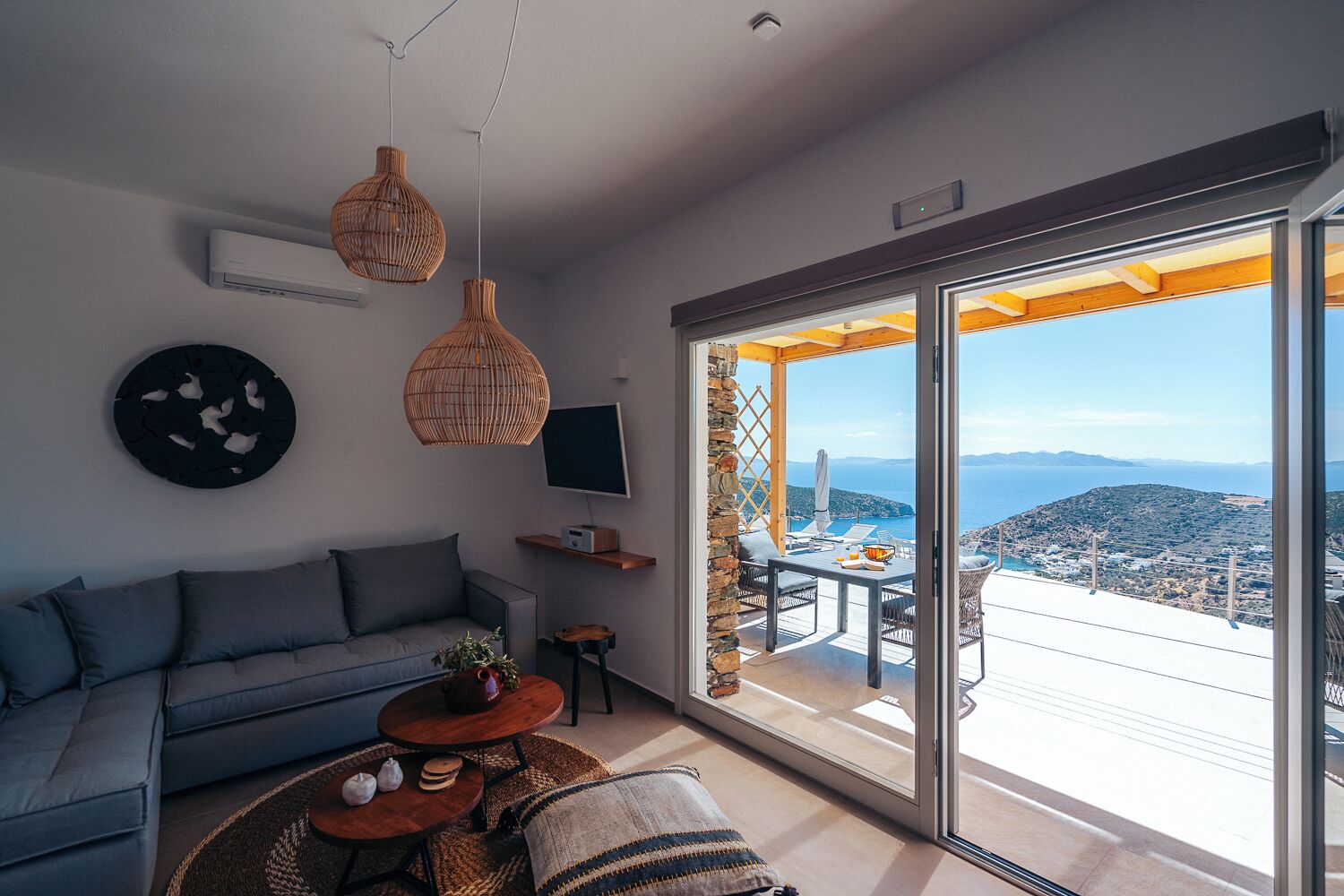 Un moderno salón con sofá, mesa de centro y televisor. Hay un balcón con vista al mar y a las colinas.