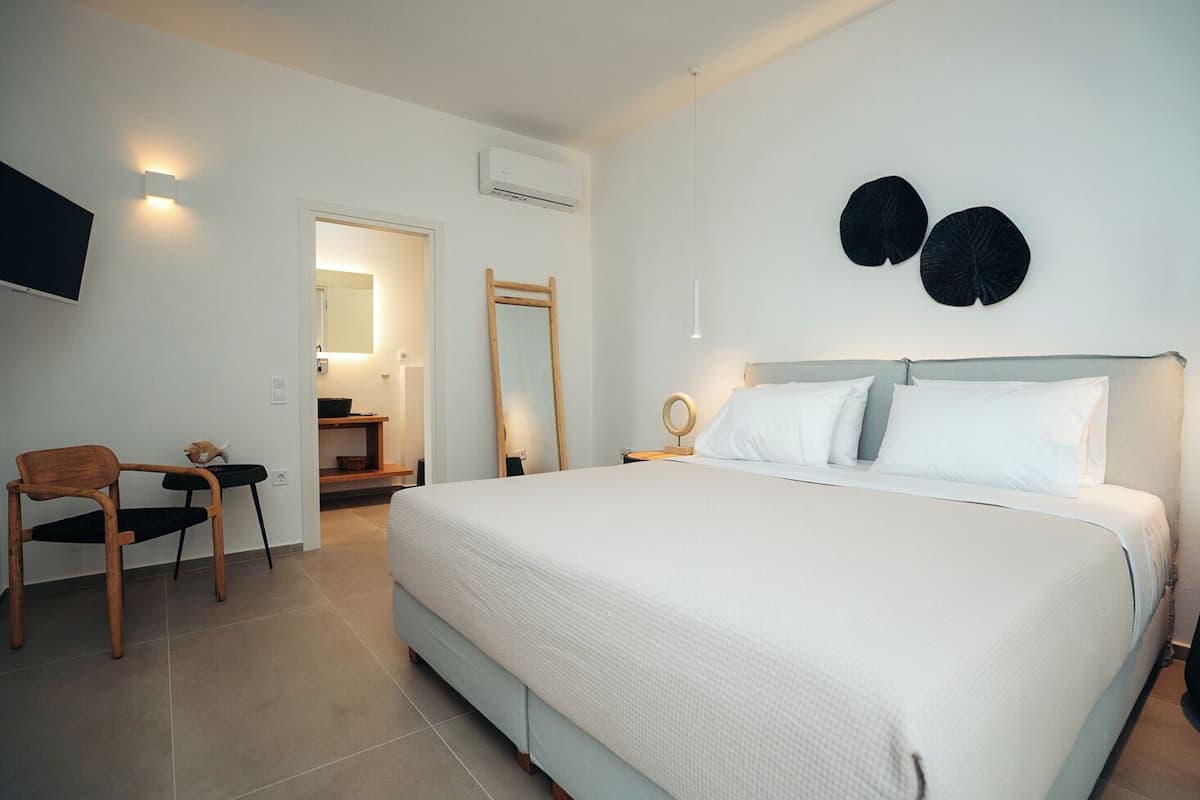 Quarto com cama grande, cadeira de madeira, mesa pequena, espelho e televisão na parede.
