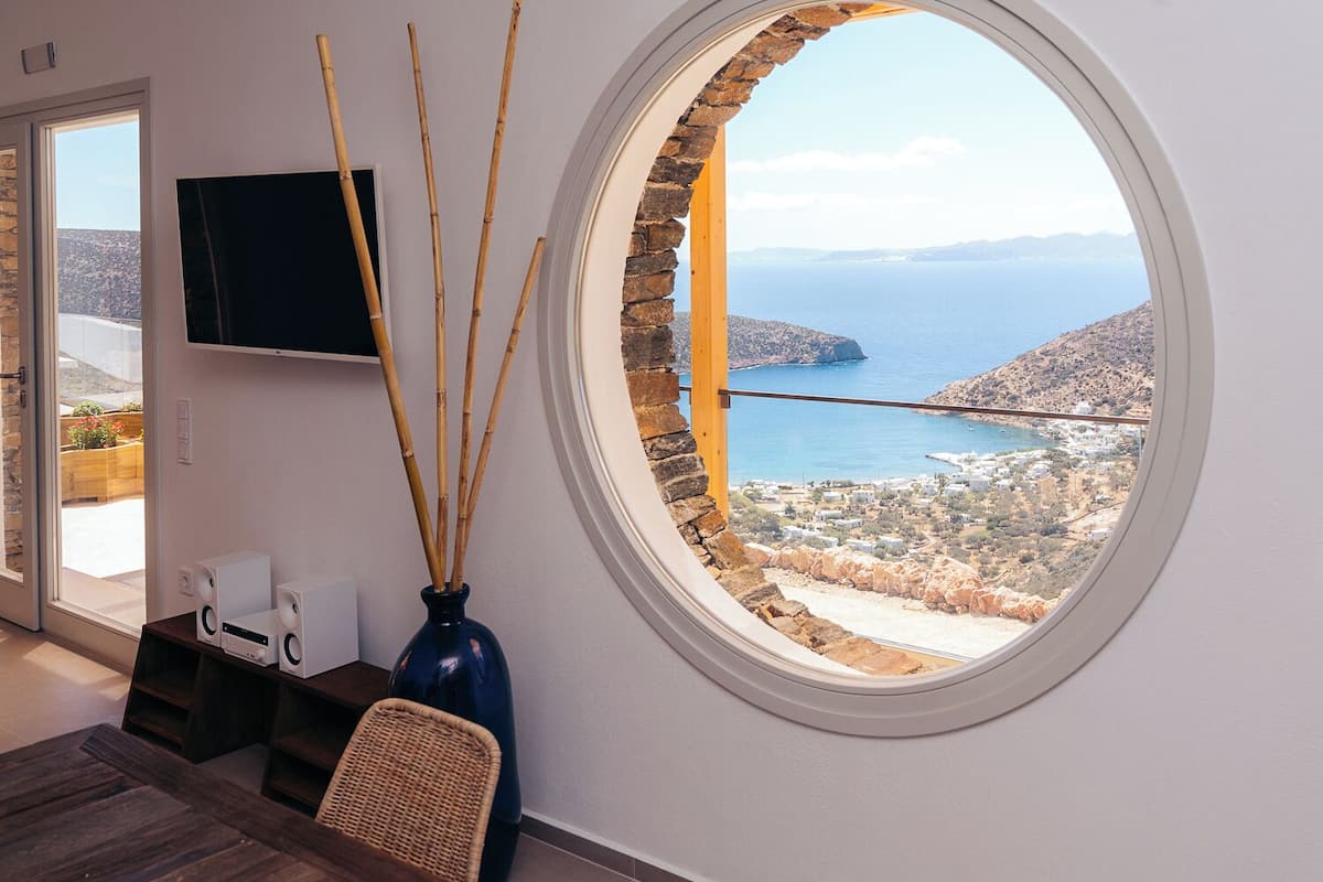 Quarto com uma grande janela redonda que oferece vista para a paisagem costeira, uma televisão de tela plana na parede e um vaso azul escuro com raminhos de junco.