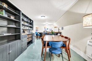 A dining area with a wooden table and blue chairs, a bookshelf, and a kitchen in the background.