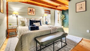 Une chambre avec un plafond en bois, un lit avec une banquette, une table de chevet avec une lampe et un tableau encadré au mur.
