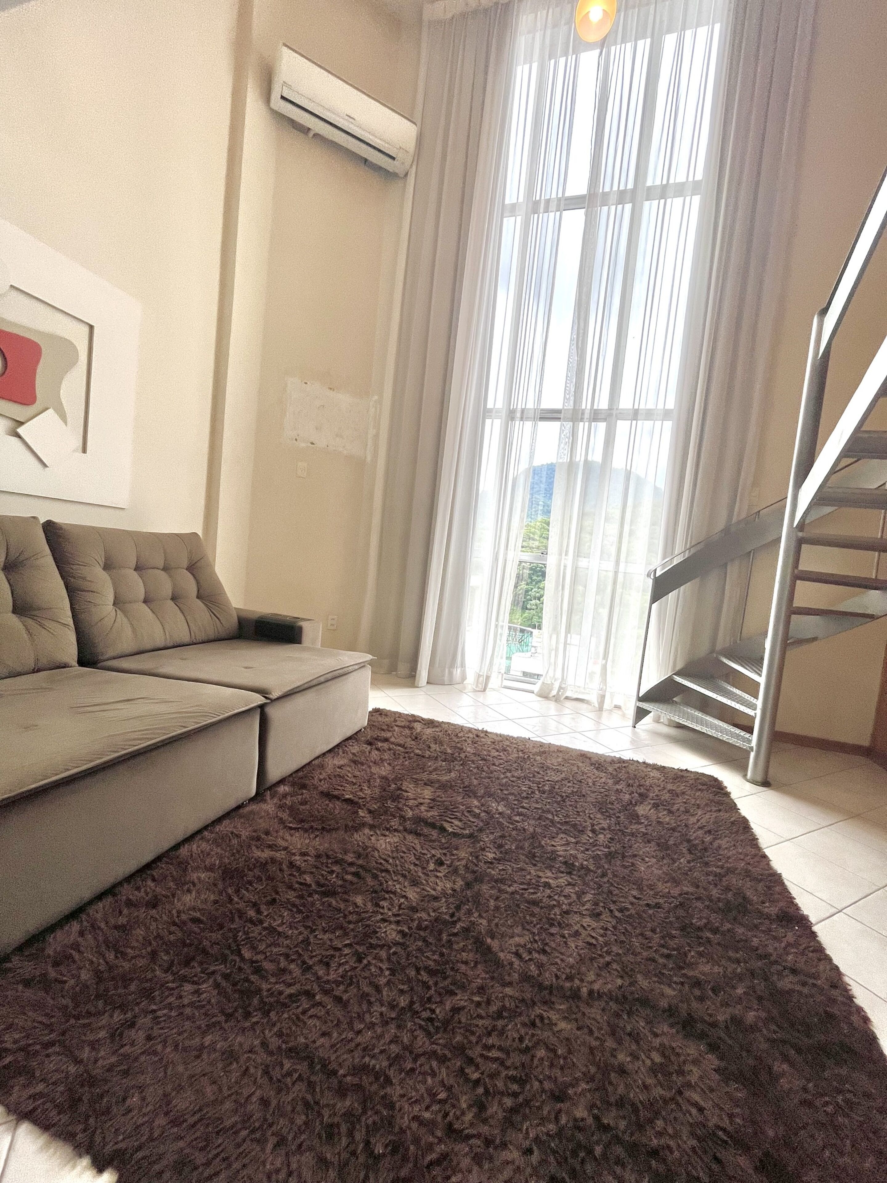 Una sala de estar con un sofá modular, un alfombra mullida oscura, una escalera y un ventanal con cortinas traslúcidas.