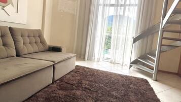 Una sala de estar con un sofá modular, un alfombra mullida oscura, una escalera y un ventanal con cortinas traslúcidas.