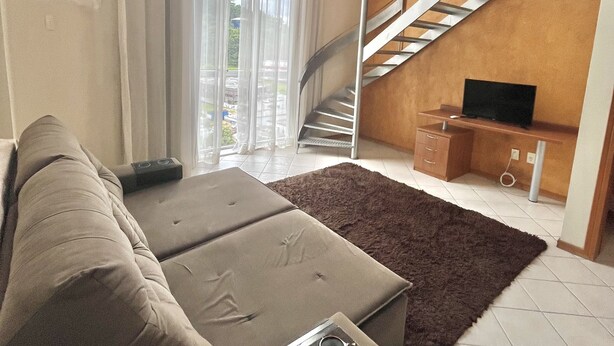 A living room with a sofa, a brown rug, a TV, and a staircase. - Saint Sebastian Flat 602Duplex no Centro (Jaragua do Sul)
